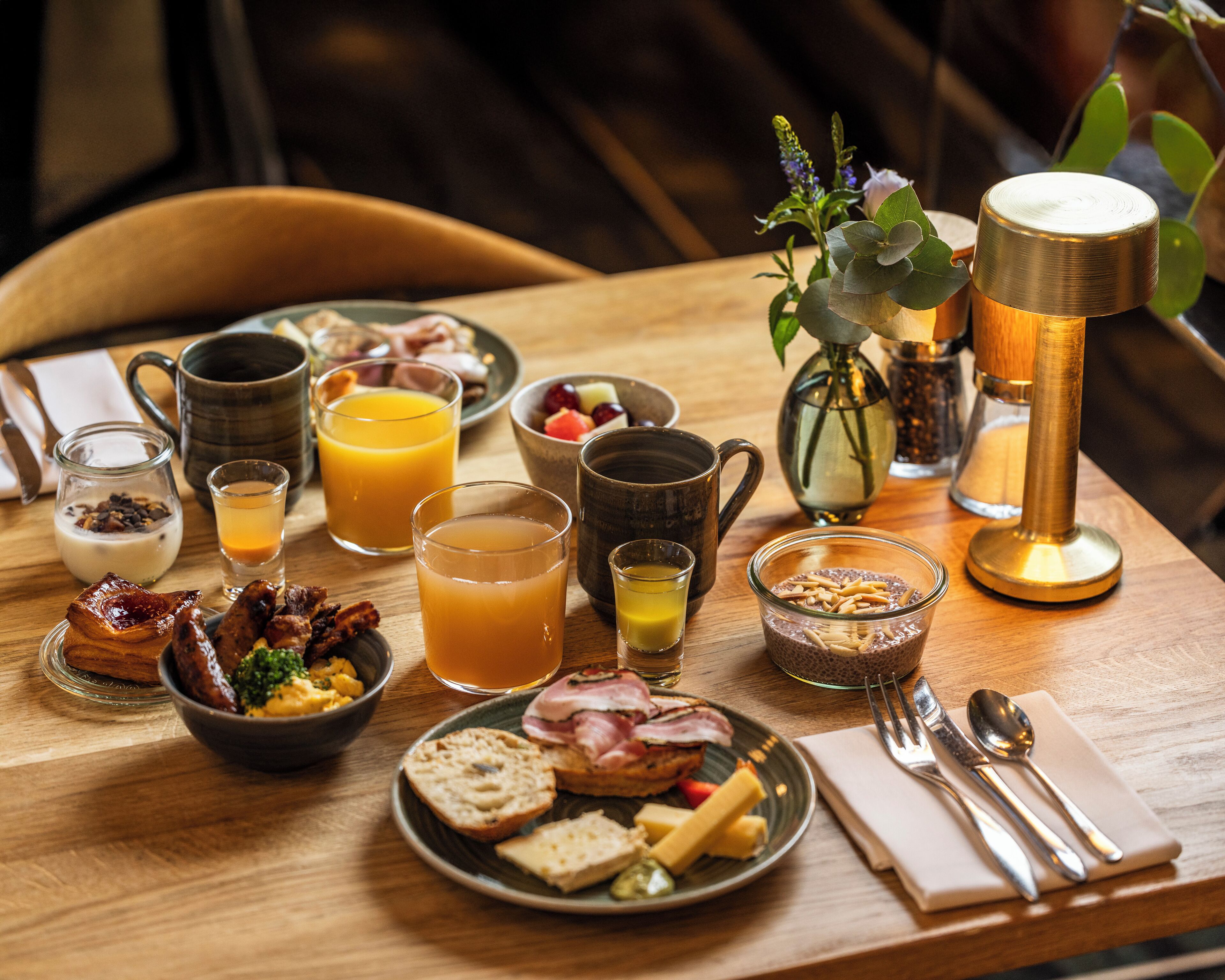 Een ontbijtsetting met diverse etenswaren, drankjes en een koffiezetapparaat op een houten tafel.