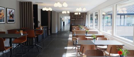 Ein modernes Restaurant mit einer langen Theke, mehreren Tischen und Stühlen. Es gibt Deckenleuchten und große Fenster.
