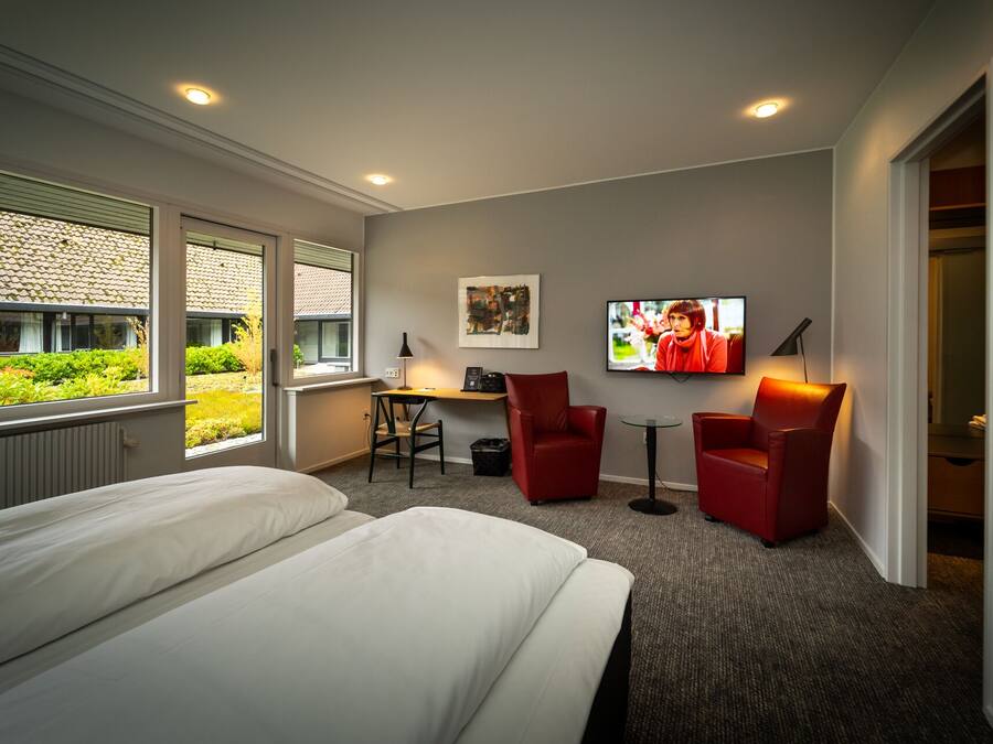 A hotel room with a bed, two red chairs, a desk, a TV, and a window with a view of greenery.