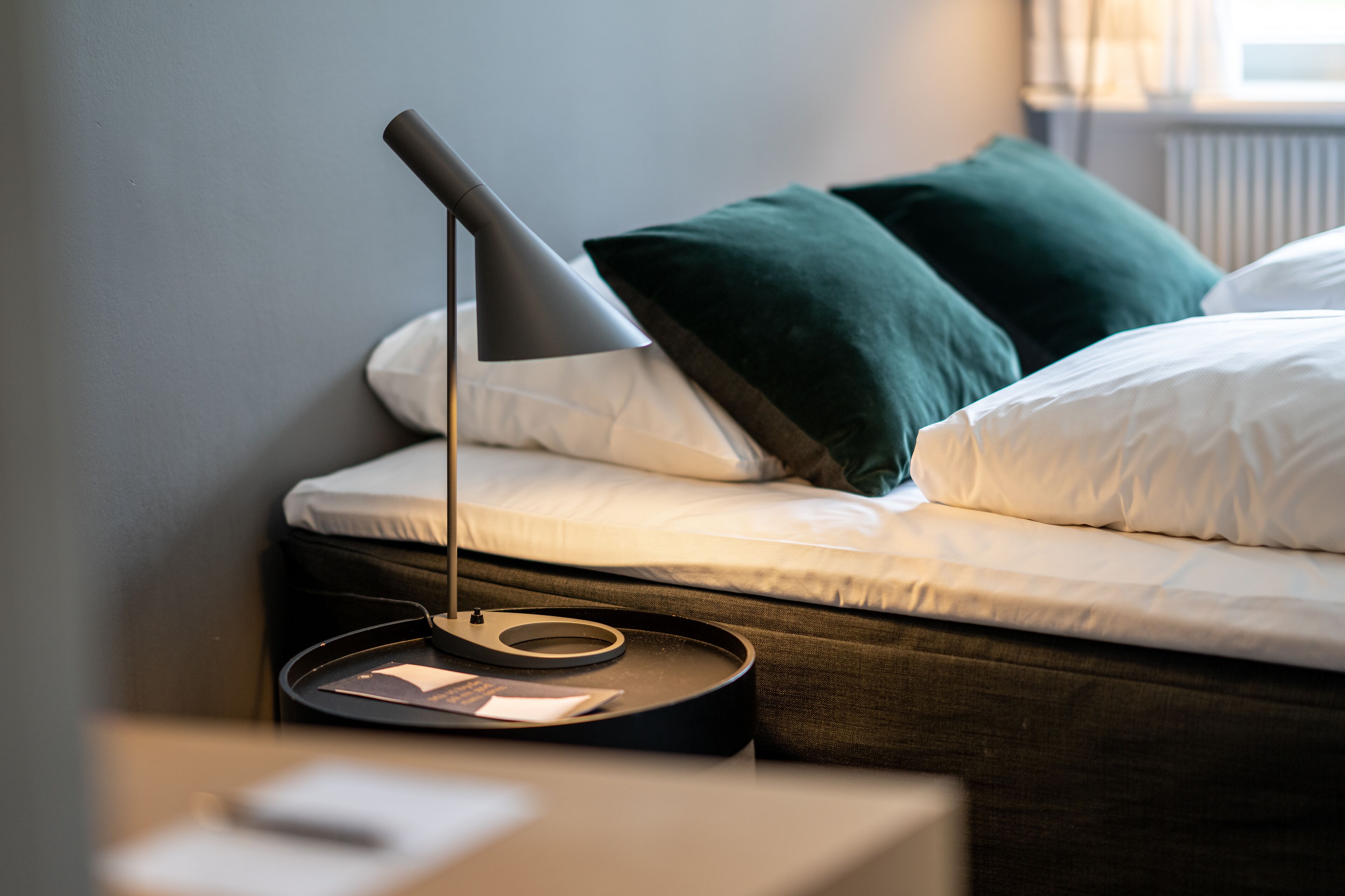 En pænt redt seng med hvide sengetøj og to grønne puder, en moderne lampe på et natbord, og et lille natbord med en bog og en fjernbetjening.