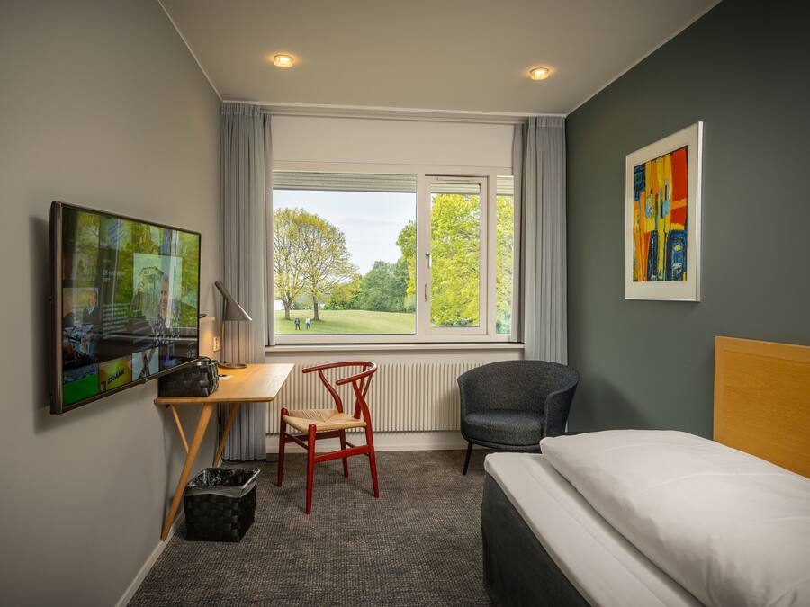 Hotelkamer met een bed, bureau, stoel, televisie en raam met uitzicht op bomen.
