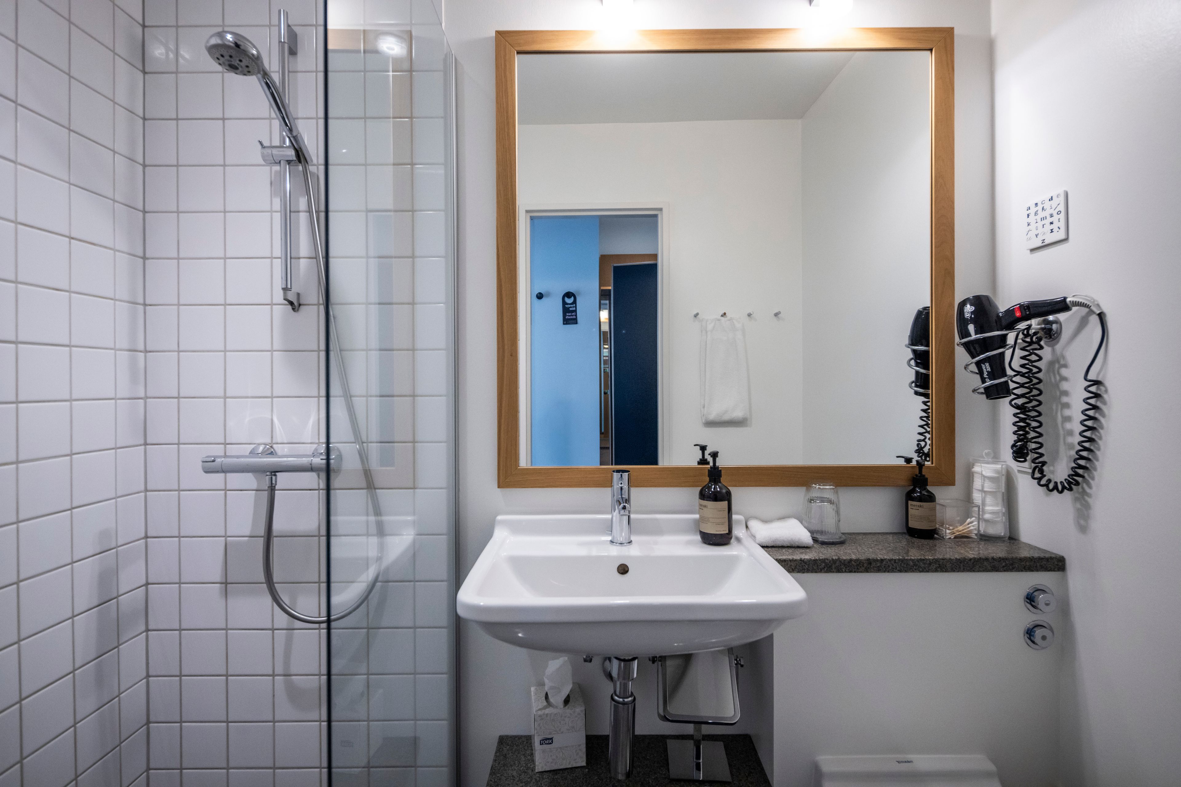 Un baño con ducha, lavamanos, espejo, toallero y secador de pelo.