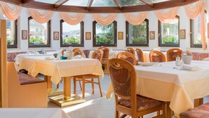Un comedor con varias mesas y sillas, amplias ventanas con cortinas y cuadros en las paredes.