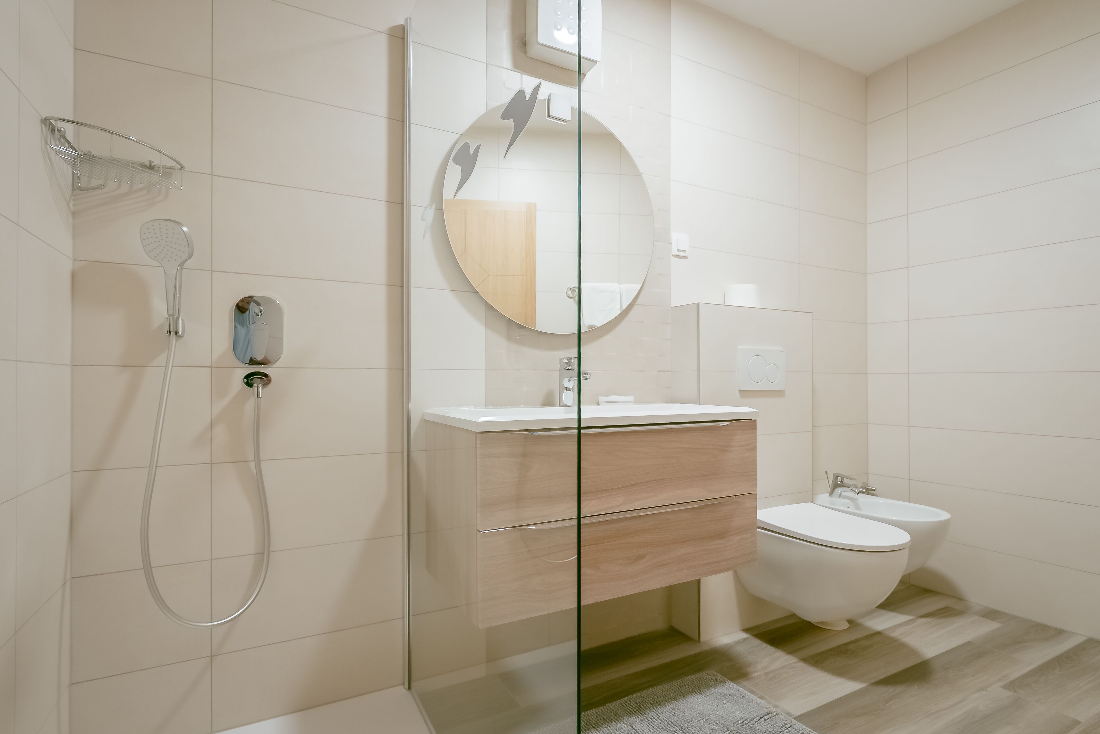 A modern bathroom with a glass shower enclosure, a round mirror, a wooden vanity, and a bidet.