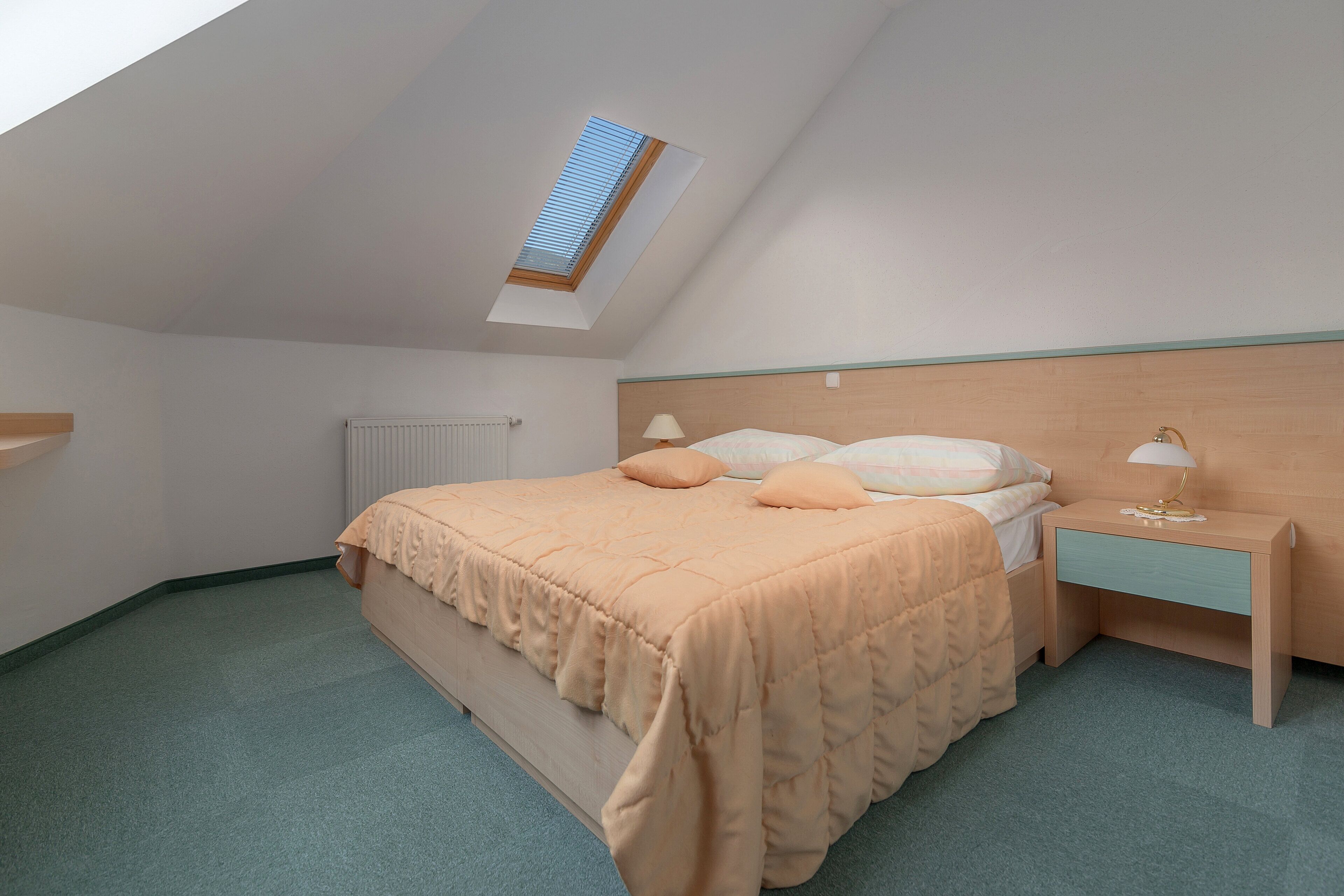 A bedroom with a sloped ceiling, a skylight, a bed with a headboard, a bedside table with a lamp, and a radiator.