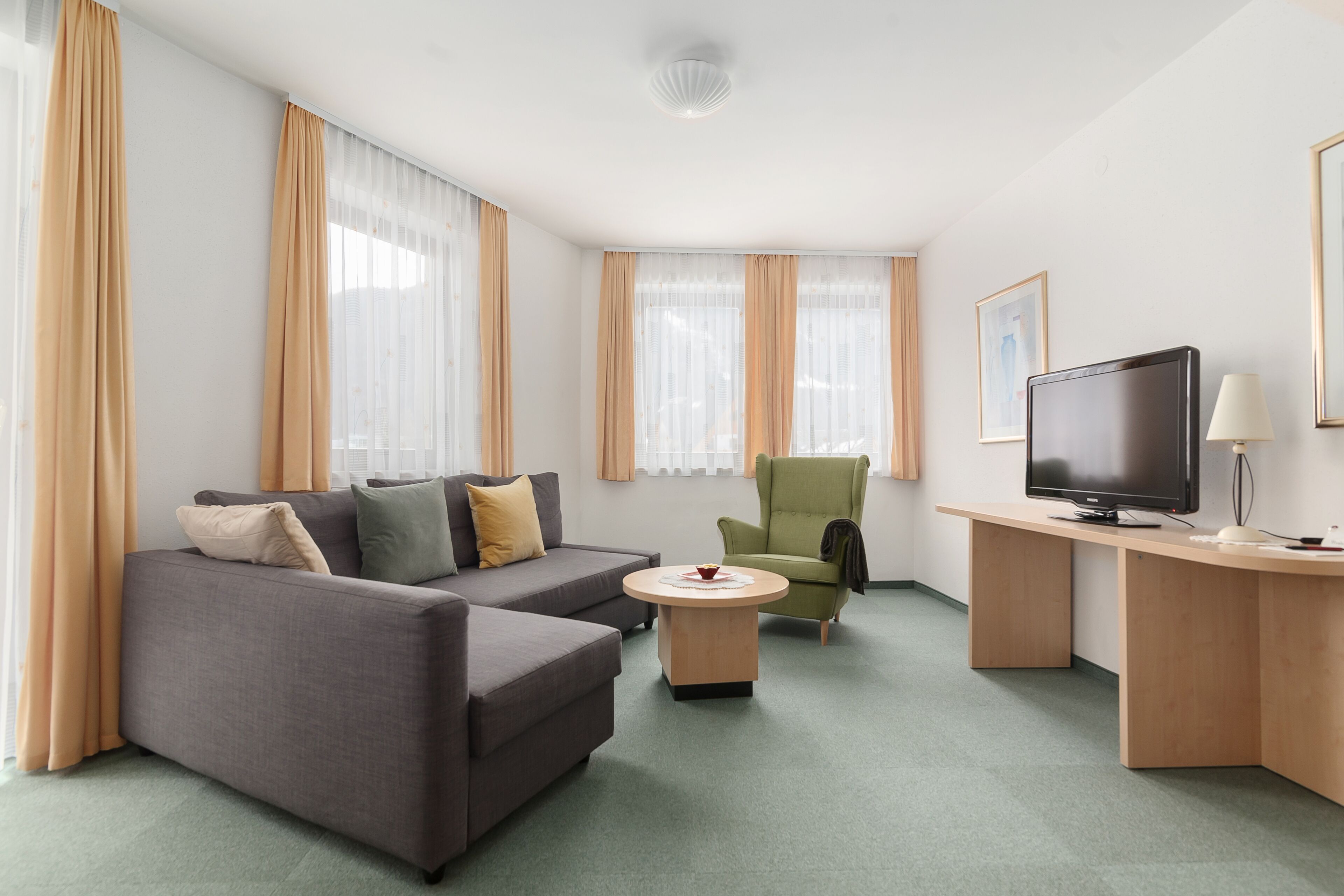 Una habitación de hotel moderna con un sofá gris, un sillón verde, un escritorio de madera con televisor y una pequeña mesa redonda.