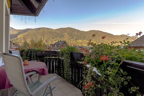 Un patio con mesa y sillas, rodeado de follaje y flores, con vistas a un paisaje montañoso.