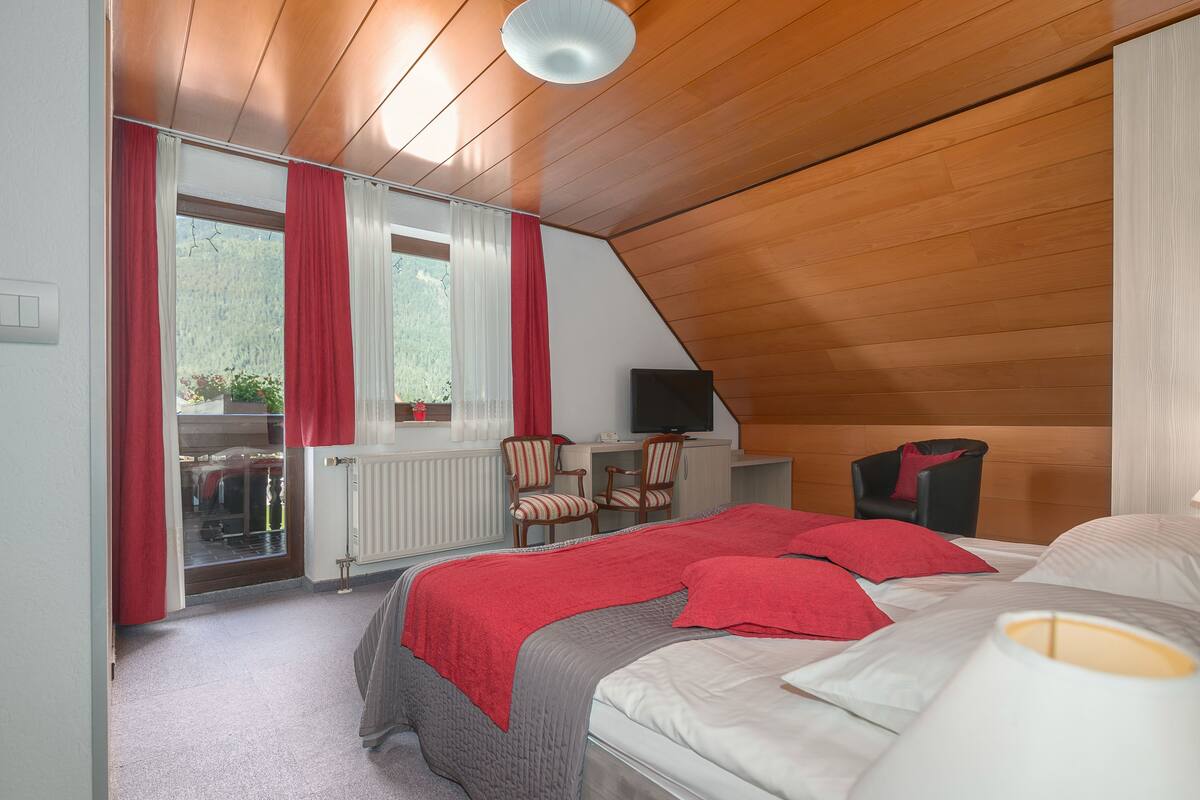 A bedroom with a sloped ceiling, a bed with red and white bedding, a desk with a chair, and a television.