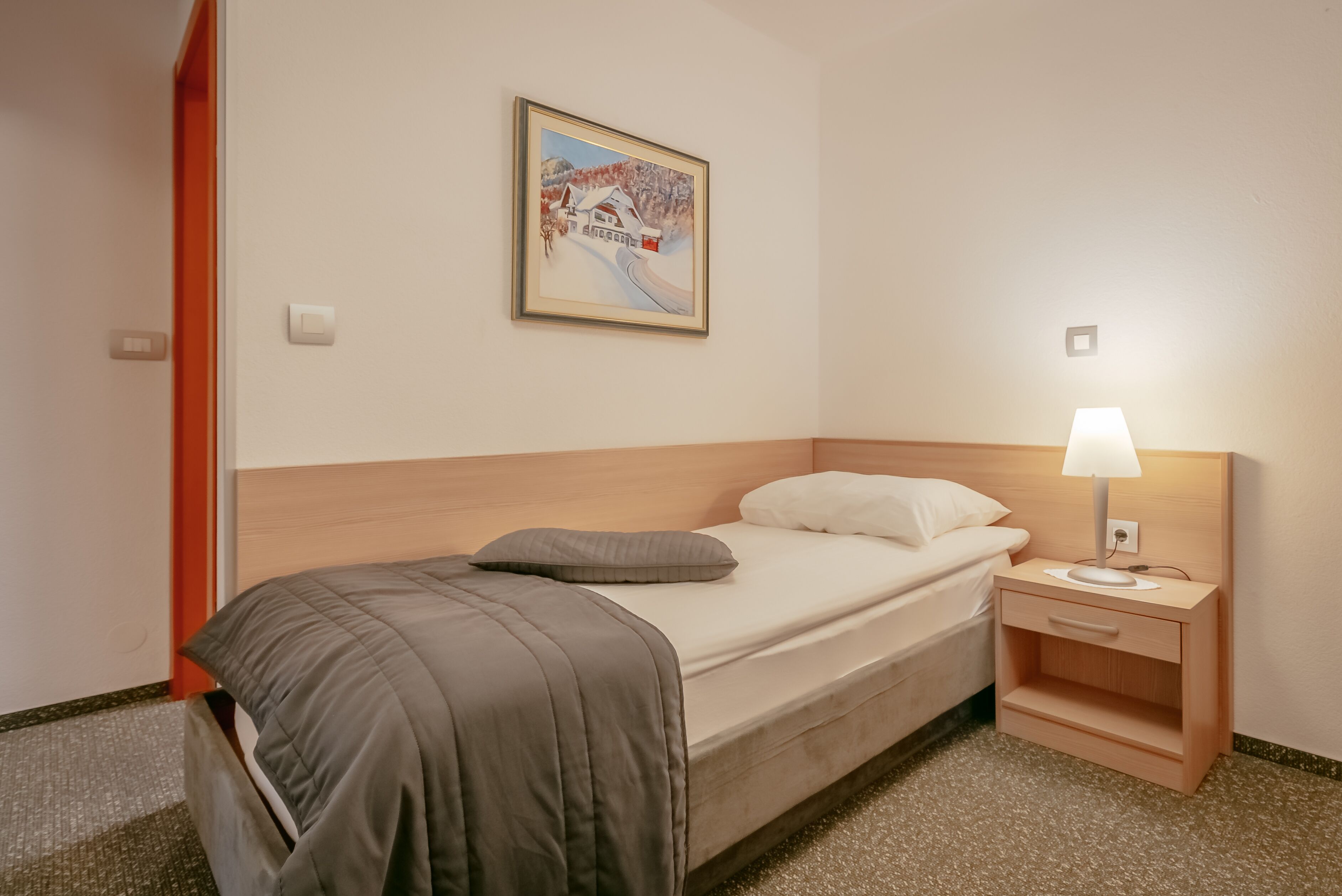 Una cama bien hecha con un edredón gris, una almohada blanca y una mesita de noche de madera con una lámpara.