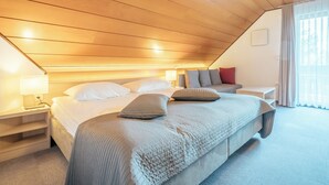 Un dormitorio con techo inclinado, una cama grande con manta acolchada, dos mesitas de noche con lámparas, una mesita auxiliar y una ventana con cortinas.
