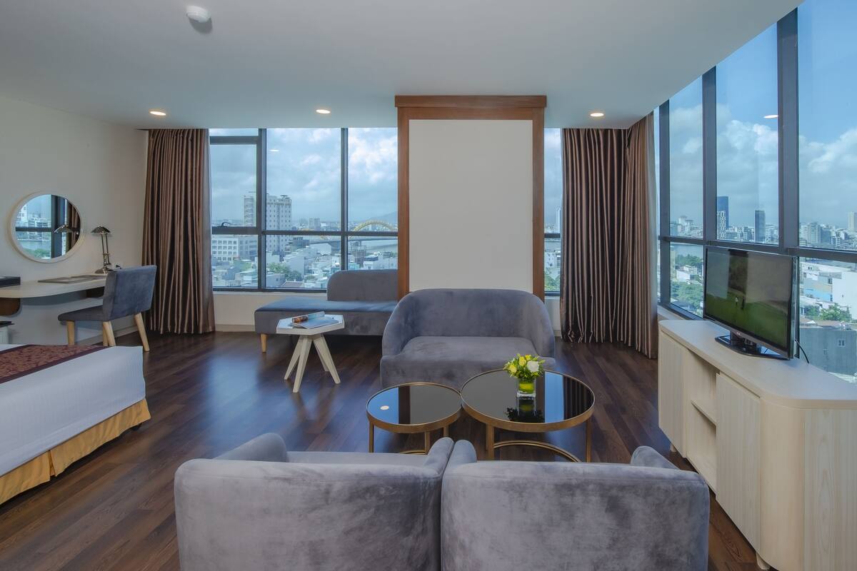 A modern hotel room with a large window, a sofa, a chair, a desk, and a television.