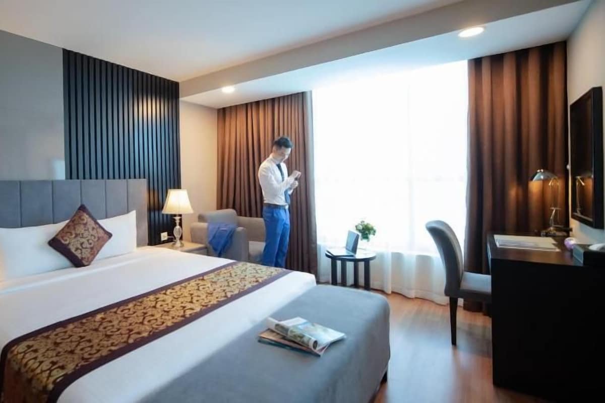Ein Hotelzimmer mit Bett, Schreibtisch, Stuhl, Lampe und einem fenster mit Vorhängen.