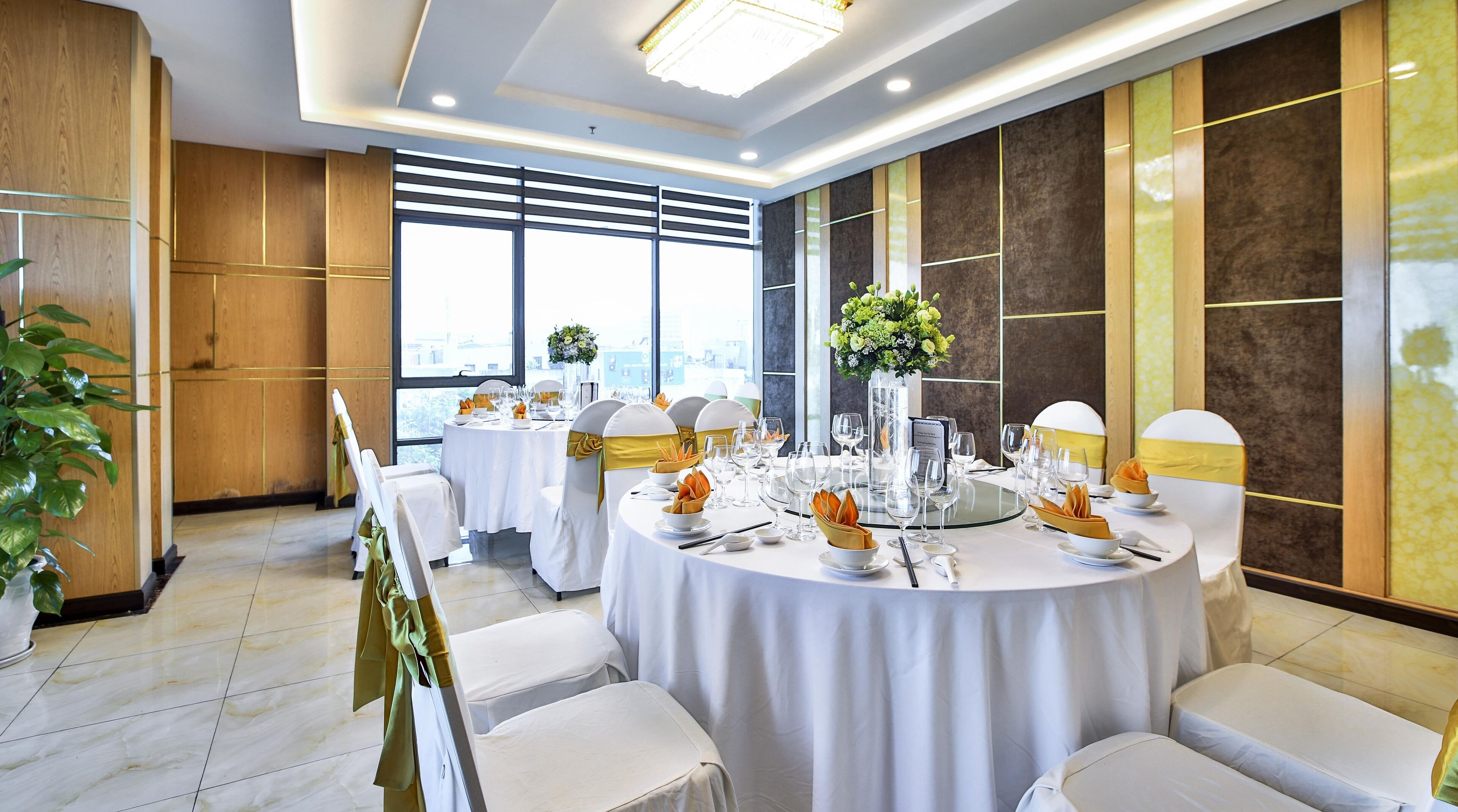 Une salle de banquet bien éclairée, avec des tables rondes dressées pour un événement formel, agrémentée de nappes blanches, de rubans dorés et de compositions florales.