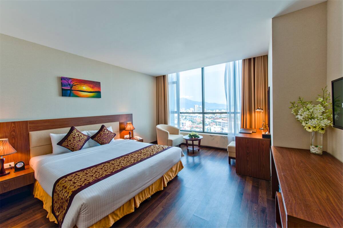 A hotel room with a large bed, a desk, a chair, a TV, and a window with a city view.