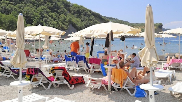A beach with rows of sun loungers and umbrellas, people swimming, and a forested hill in the background. - San Simon Resort Dependences (Izola)