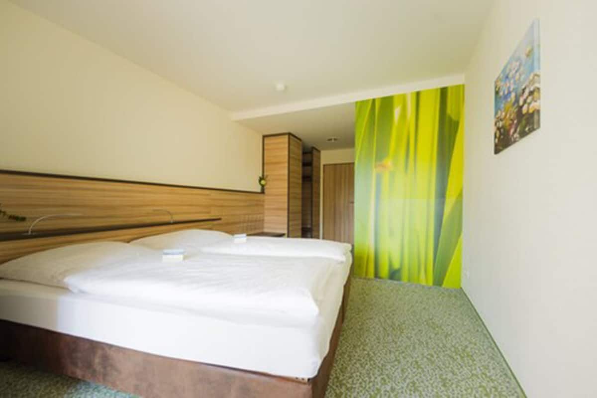 Une chambre d’hôtel avec une tête de lit en bois, un lit recouvert de draps blancs, une moquette verte et un tableau accroché au mur.