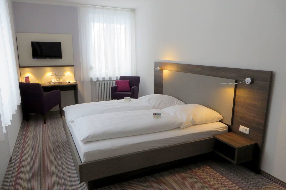 Une chambre d’hôtel comprenant un lit, un bureau avec une chaise, une télévision et une fenêtre avec des rideaux.