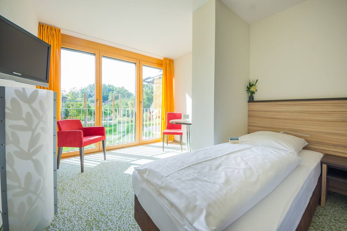 Une chambre d’hôtel avec un lit, un bureau et une chaise. Elle dispose d’un balcon avec des rideaux orange et offre une vue sur la verdure.
