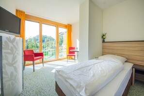 Hotellihuone, jossa on sänky, työpöytä ja tuoli. Huoneessa on parveke, jossa on oranssit verhot ja näkymä vehreään maisemaan.