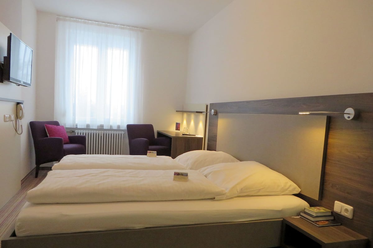 Une chambre d’hôtel avec un grand lit, deux fauteuils, un bureau et un téléviseur fixé au mur.