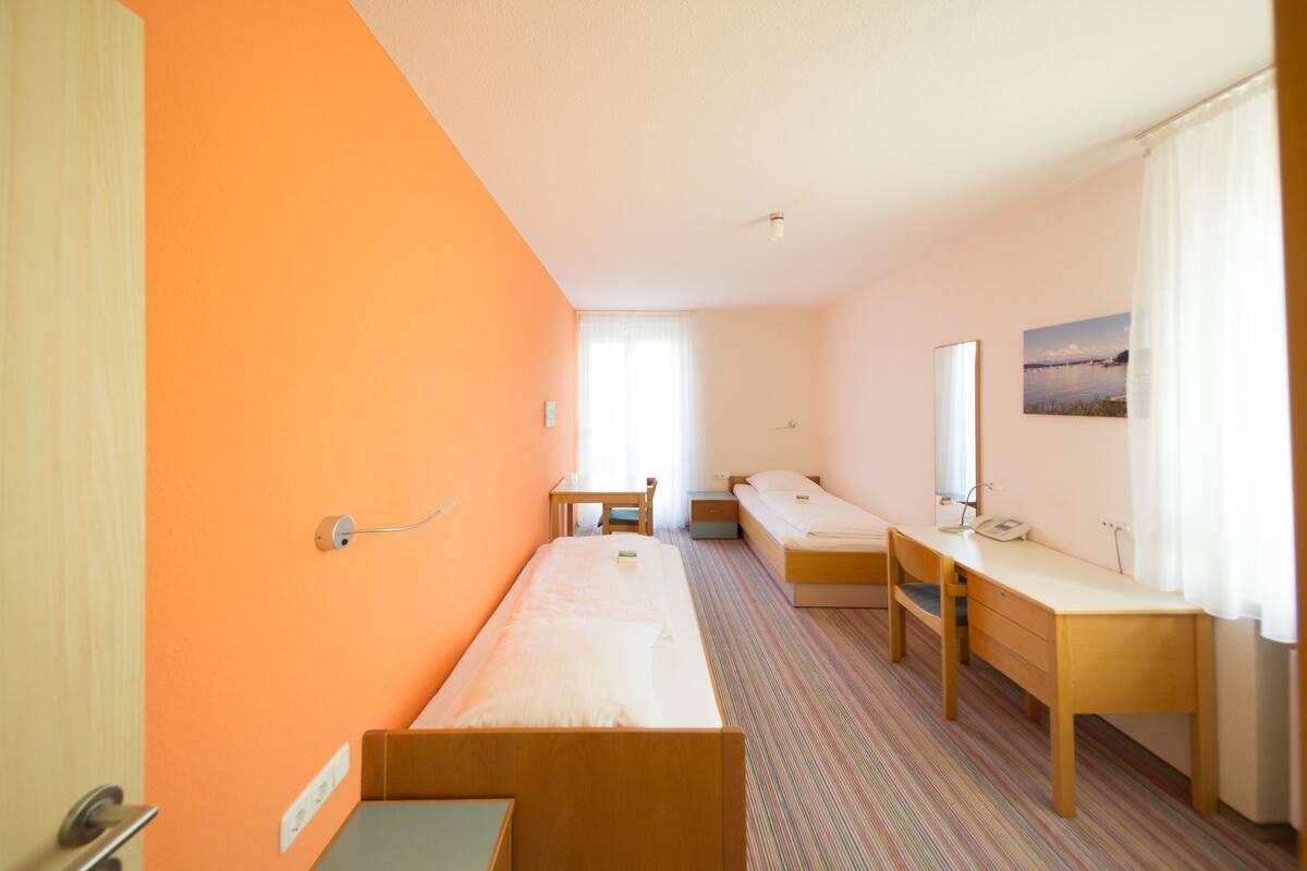 Une chambre d’hôtel avec un lit simple, un bureau, une chaise, une table de chevet et un miroir.