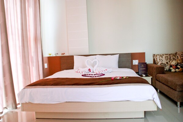 A neatly made bed with a white and brown bedspread, a decorative swan pillow, and a heart-shaped pillow. There is a brown sofa with a teddy bear and a small table with a book and a vase of flowers.