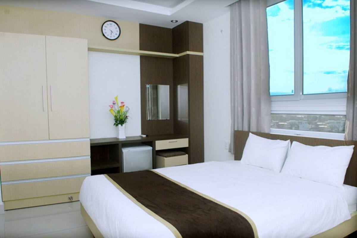 A hotel room with a bed, a desk, a wardrobe, a clock, a mirror, and a window with a view.