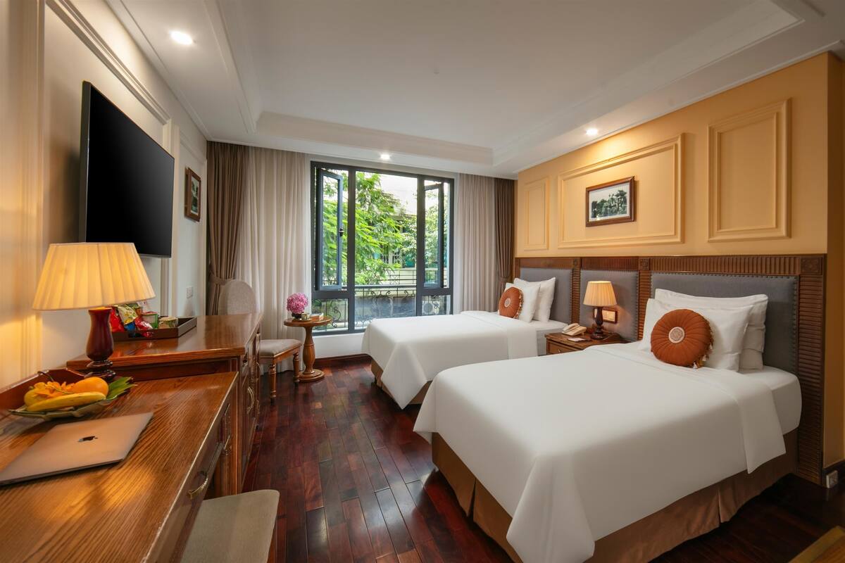 A hotel room with two beds, a desk with a laptop, a TV, and a window with a view of greenery.