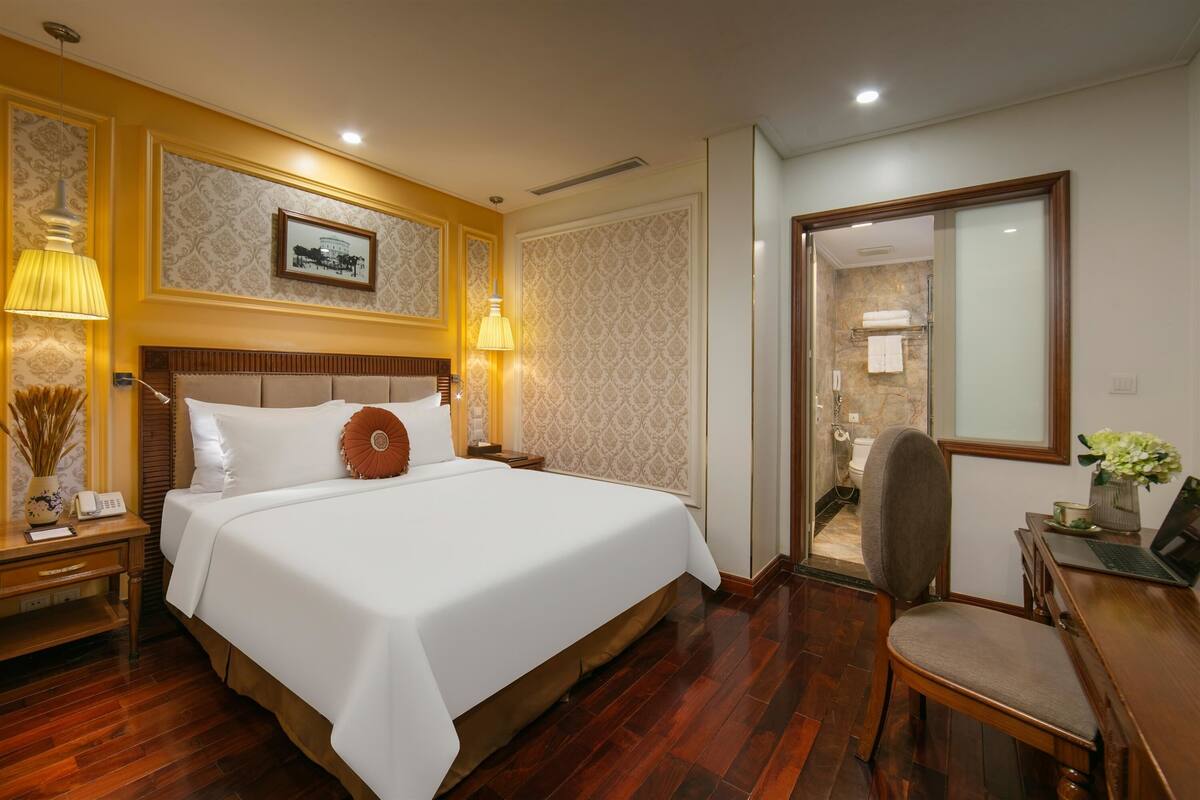 A hotel room with a large bed, bedside lamps, a desk with a chair, and a bathroom visible through an open door.