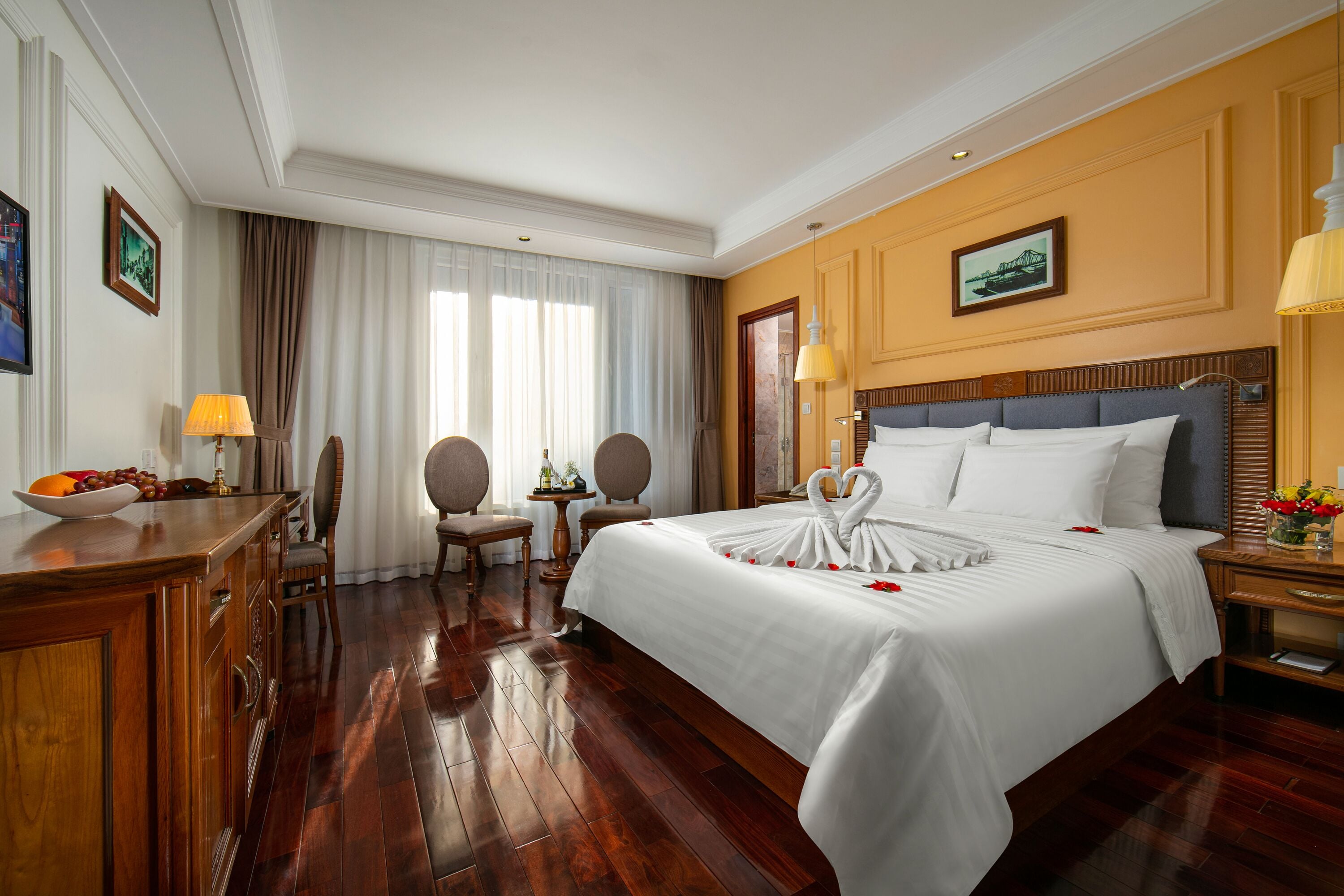 A hotel room with a large bed, wooden furniture, a desk with a lamp, and a small table with a vase of flowers.