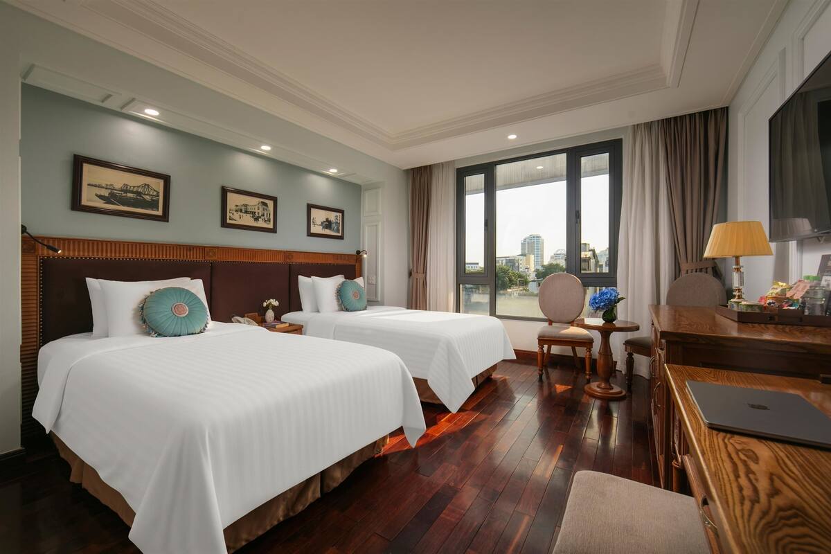 A hotel room with two beds, a desk with a laptop, a chair, and a view of the city through large windows.