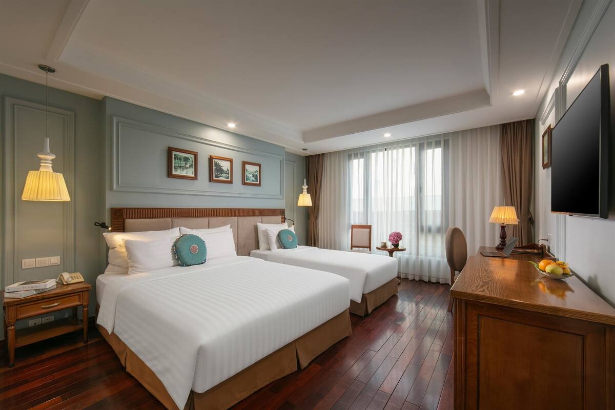 A hotel room with two beds, a desk, a TV, and a window with curtains.