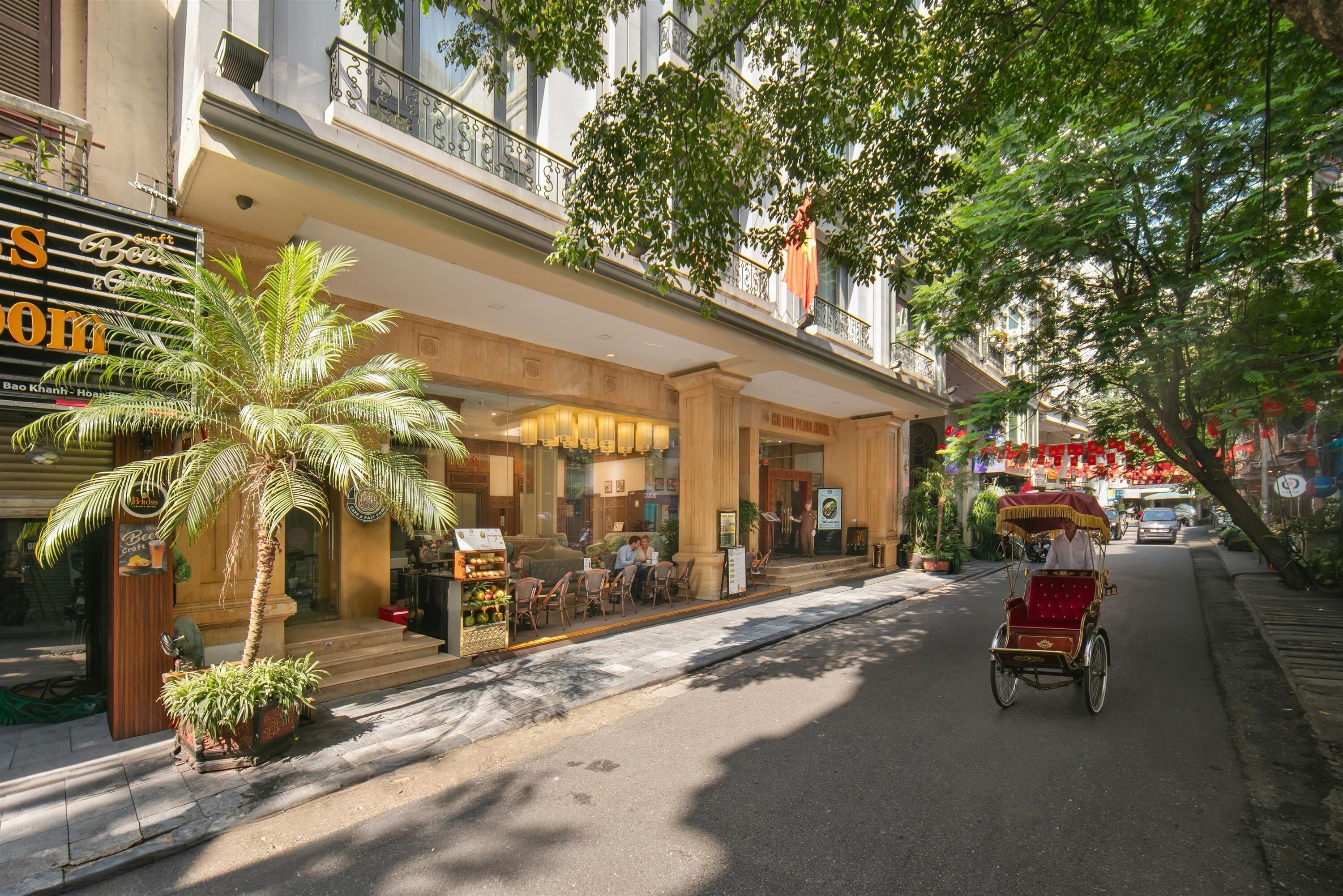 A street view of a hotel with a palm tree, outdoor seating, and a rickshaw.