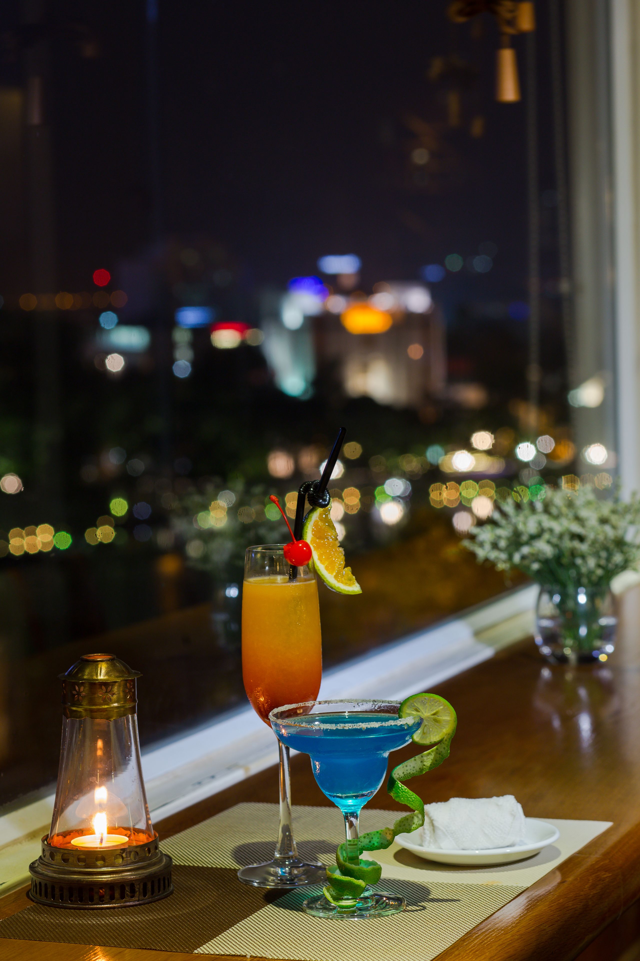 A bar setting with two drinks, a lit candle, and a view of a cityscape at night.