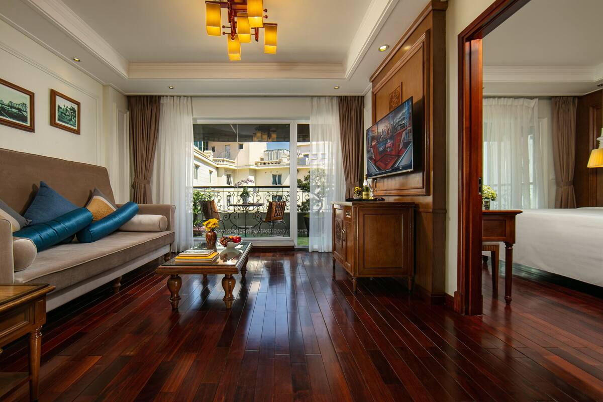 A hotel room with a sofa, a dining table with books and flowers, a TV, a bed, and a view of the outside balcony.