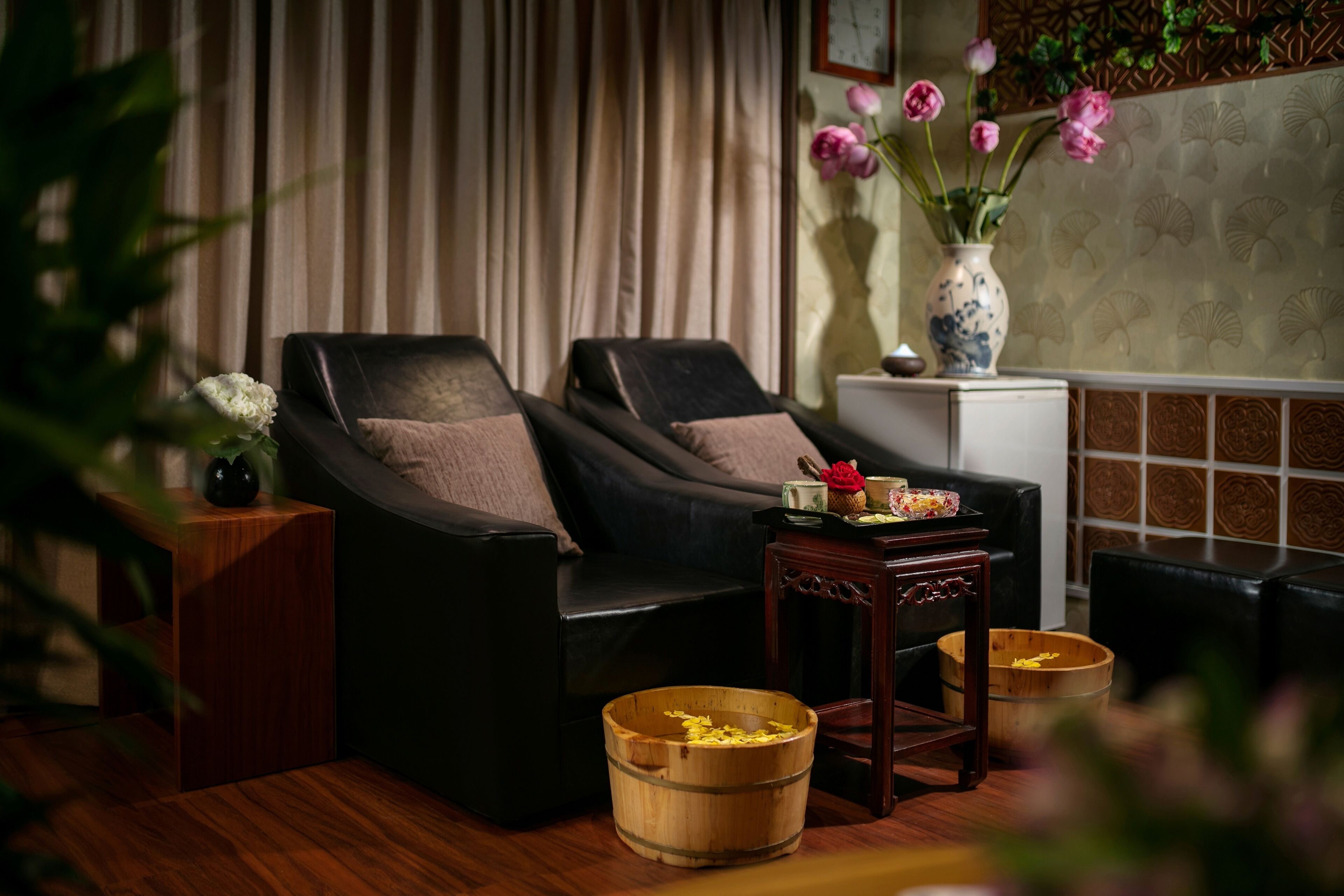 A room with two black leather sofas, a small wooden table, a vase with pink flowers, and a wooden bucket on the floor.