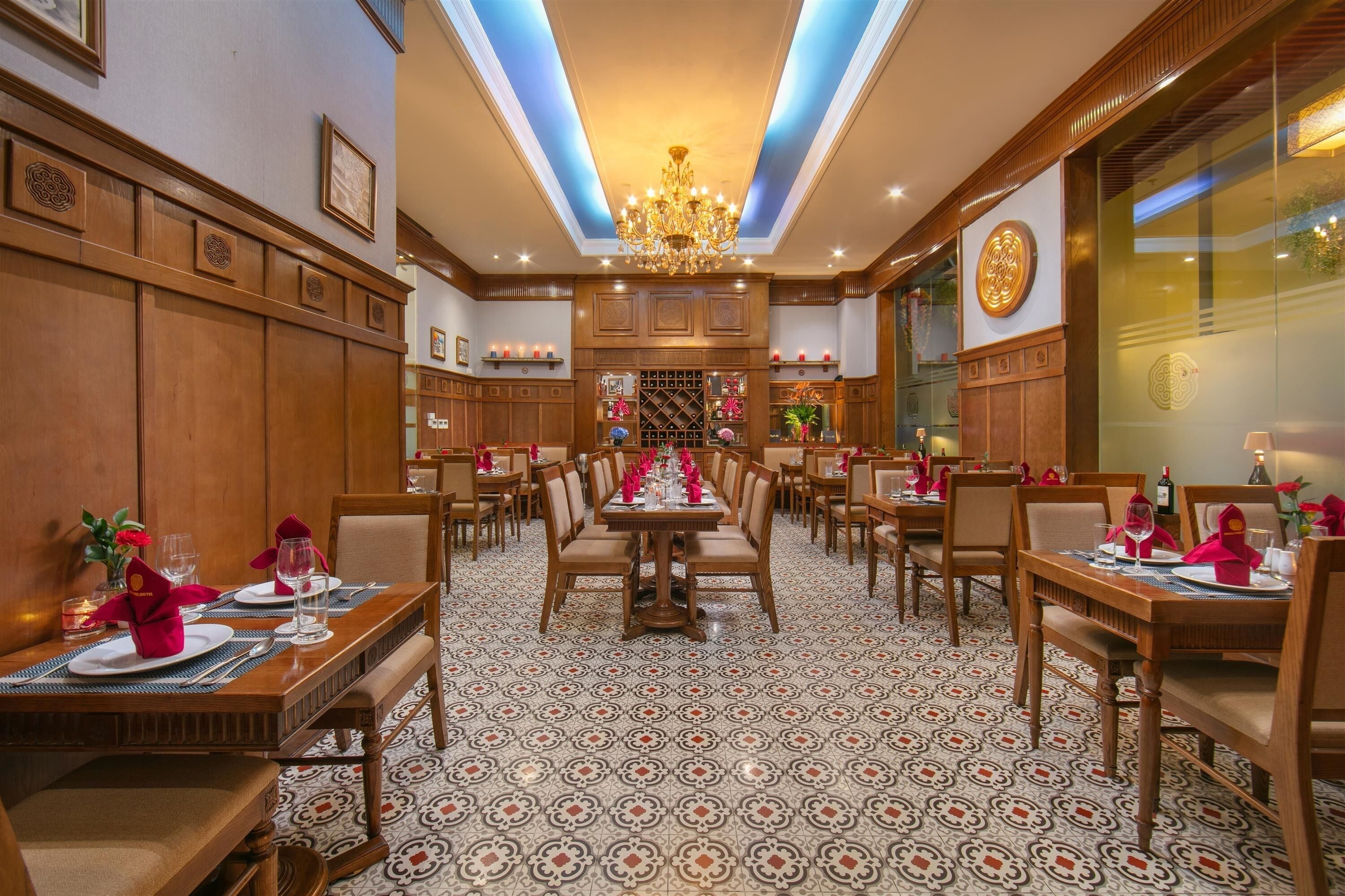 A hotel dining area with multiple tables set for dining, a chandelier, and patterned floor tiles.