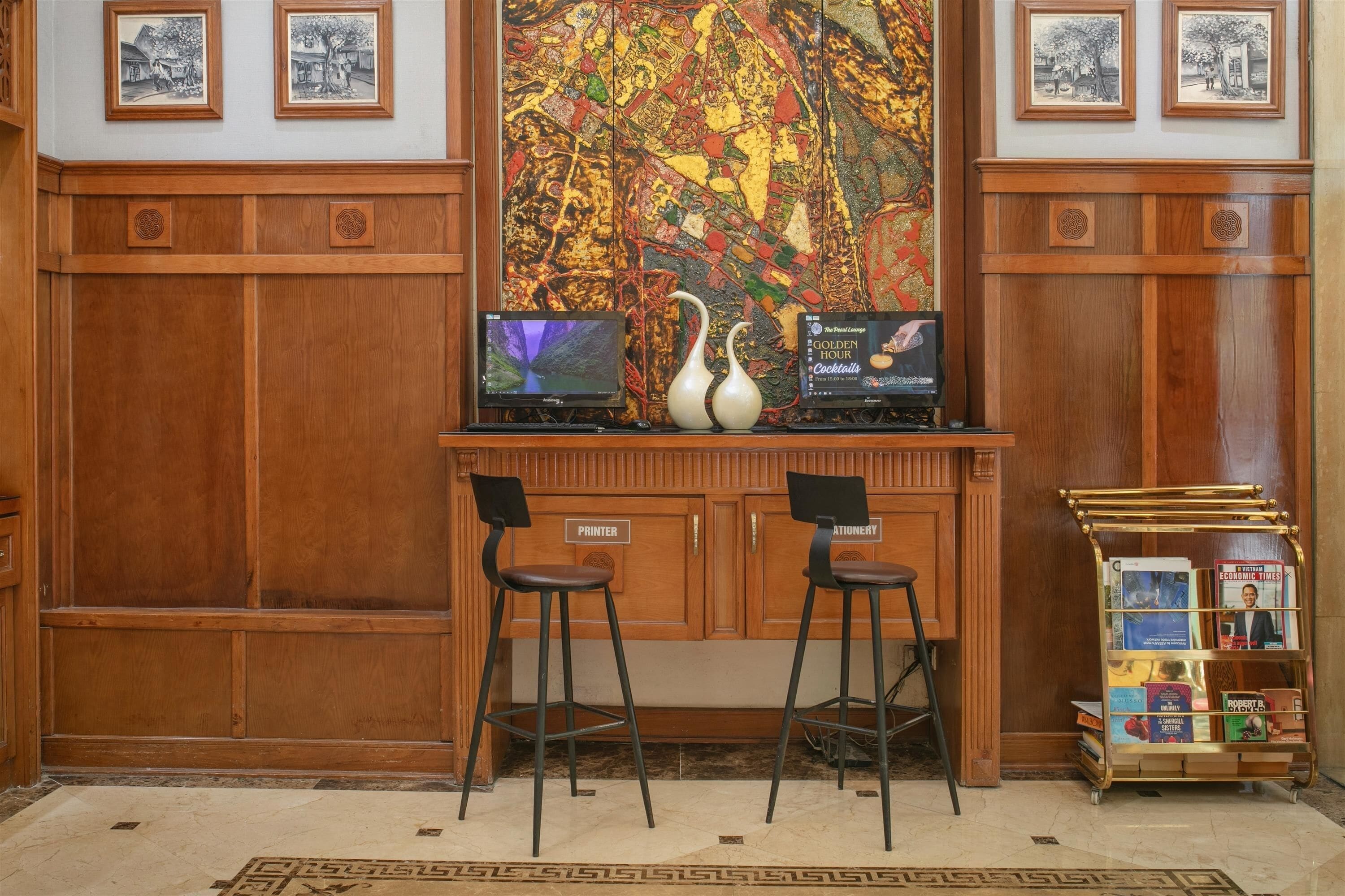 A wooden reception desk with two bar stools, a large abstract painting, and framed pictures above.