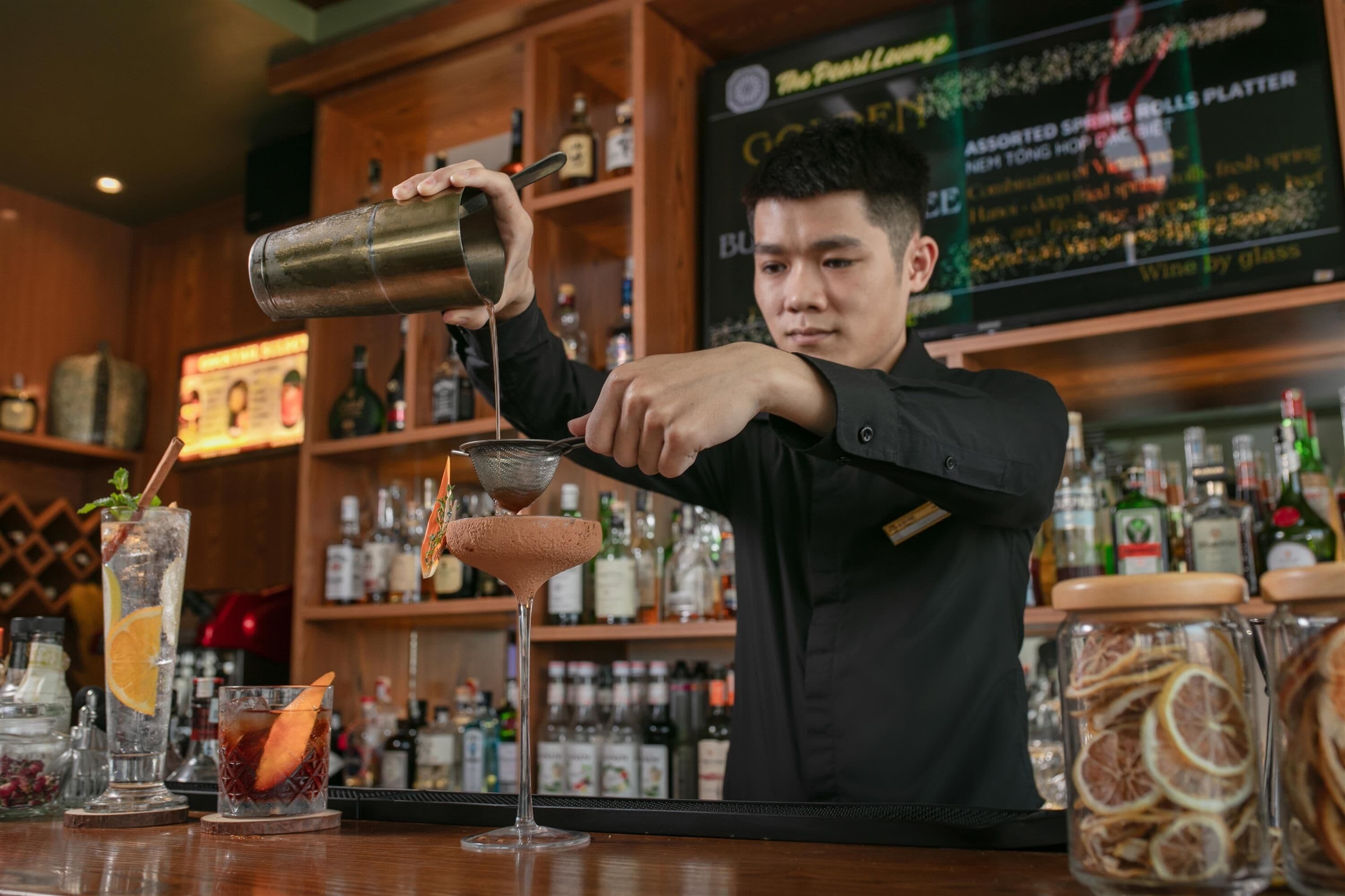 Un cantinero preparando un cóctel en la barra, con varias botellas y un menú al fondo.