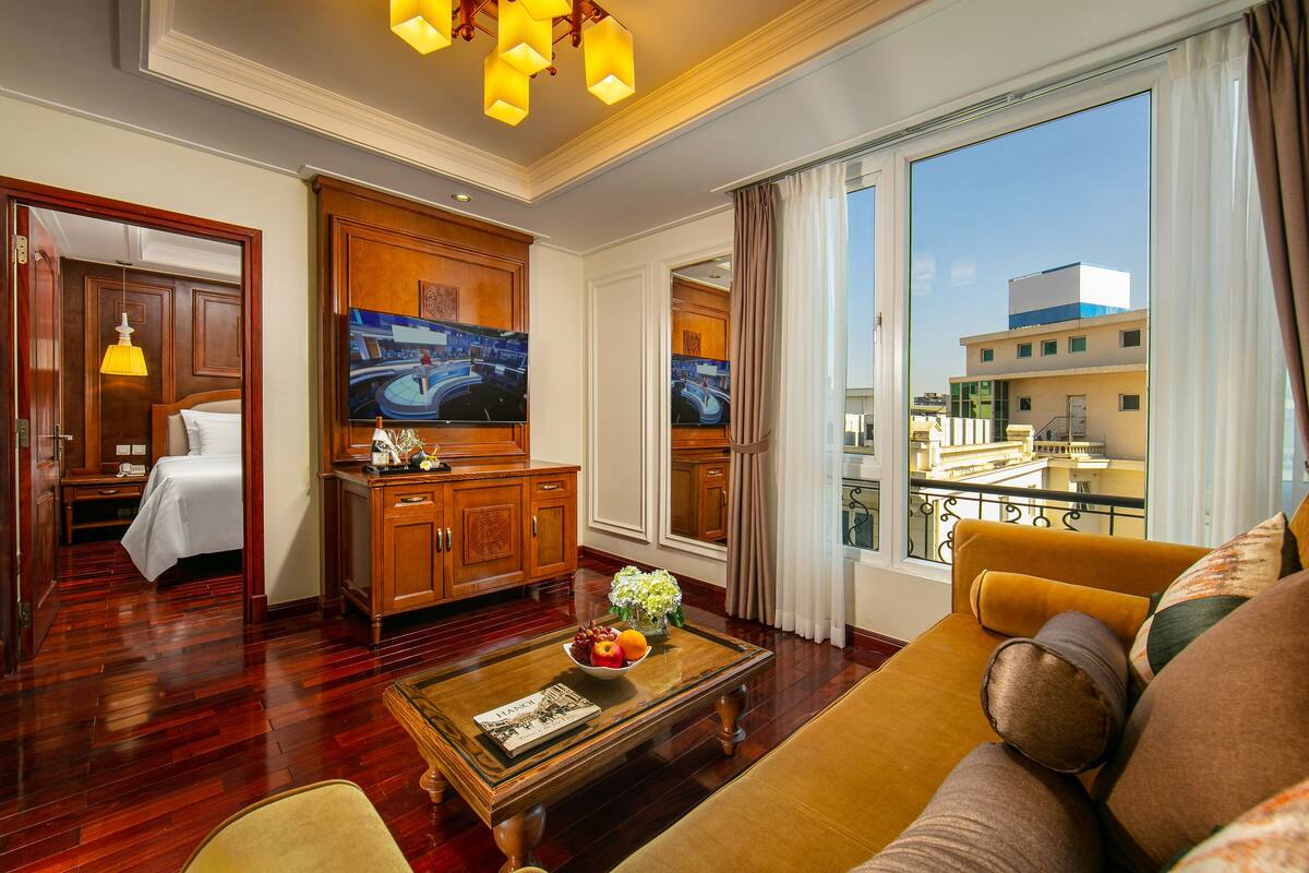 A hotel room with a large window, a sofa, a coffee table with fruit, and a television.