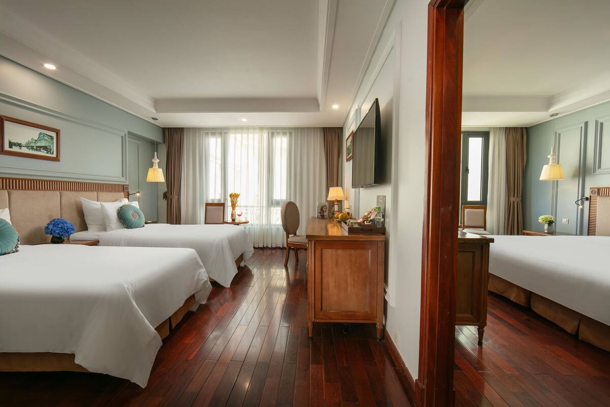 A hotel room with two beds, a wooden desk, a TV, and a large window with curtains.