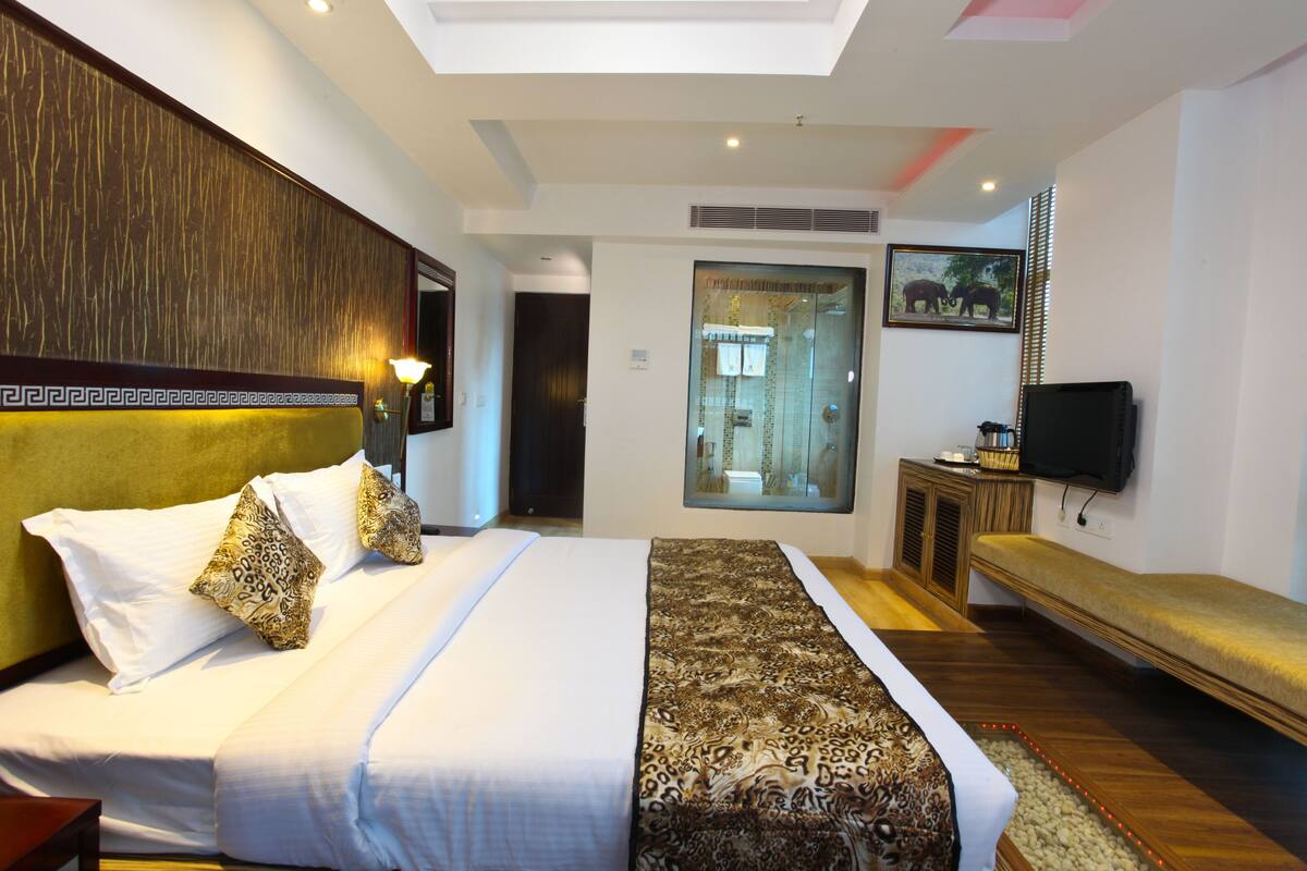 A hotel room with a large bed, a bench, a television, and a bathroom visible through a glass door.