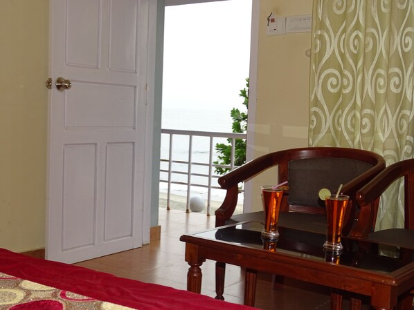 Una habitación con una mesa de madera, dos sillas, una cama con cubrecama rojo y vistas al mar a través de una puerta de balcón, además de una cortina estampada.