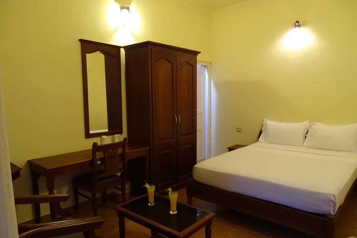 A bedroom with a bed, a wooden desk, a chair, a small table with two drinks, and a wardrobe.