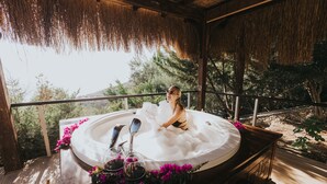 Un jacuzzi con cubierta de madera, rodeado de follaje y una estructura de techo de paja.