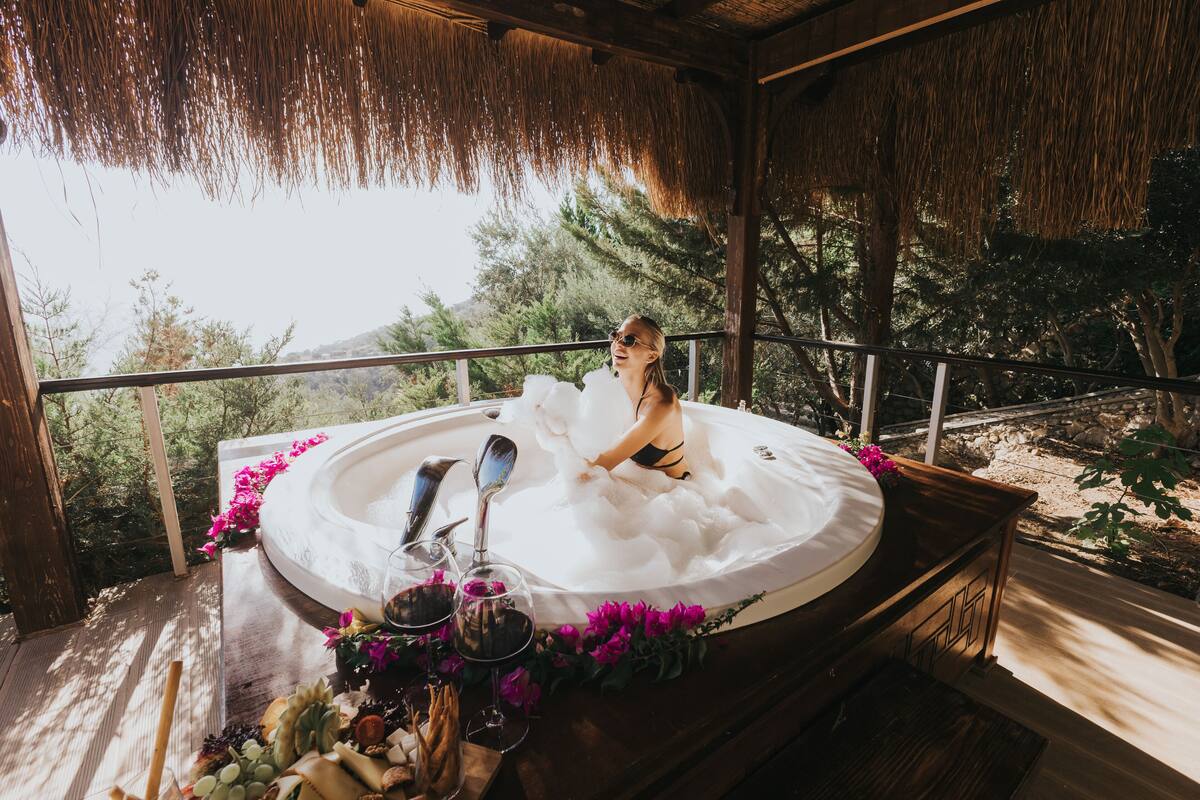 A hot tub with a wooden deck, surrounded by greenery and a thatched roof structure.