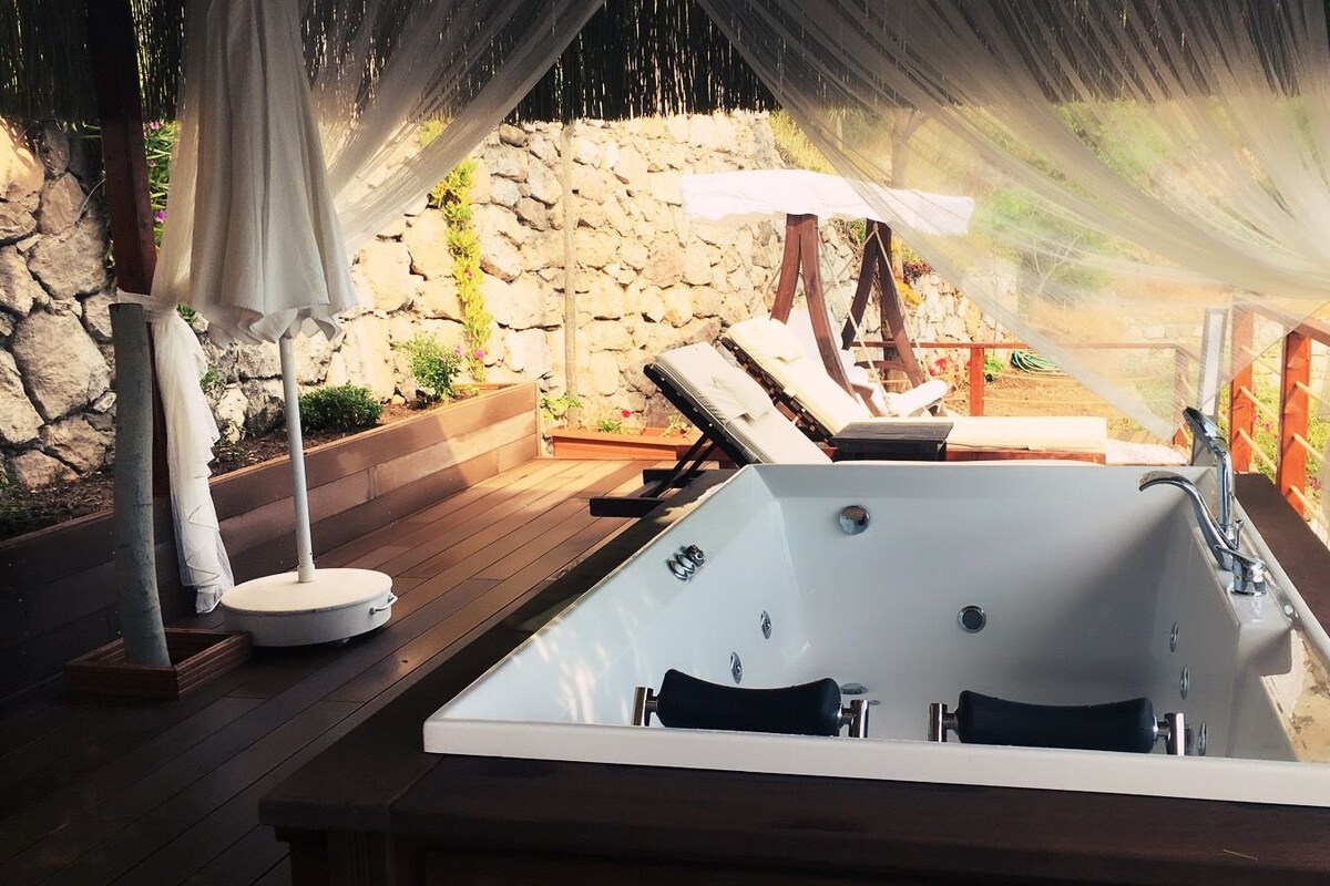 A spa area with a large bathtub, wooden deck, stone wall, and outdoor furniture.