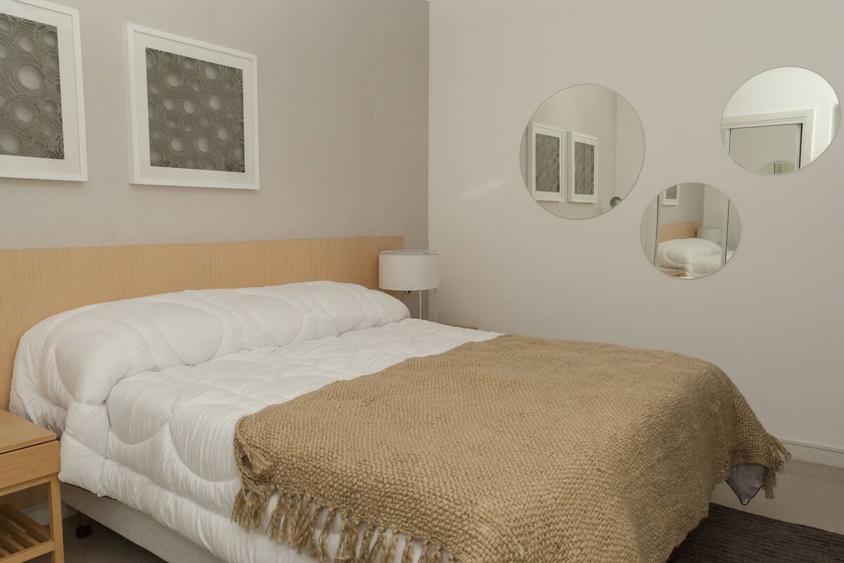 A neatly made bed with a white comforter and a beige throw blanket, a bedside table with a lamp, and three wall-mounted mirrors.