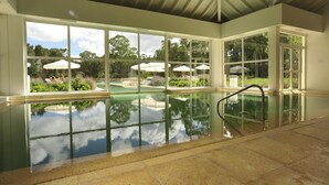 Ein großes Hallenbad mit Glaswänden, gefliestem Boden und Blick auf einen Garten mit Sonnenschirmen.