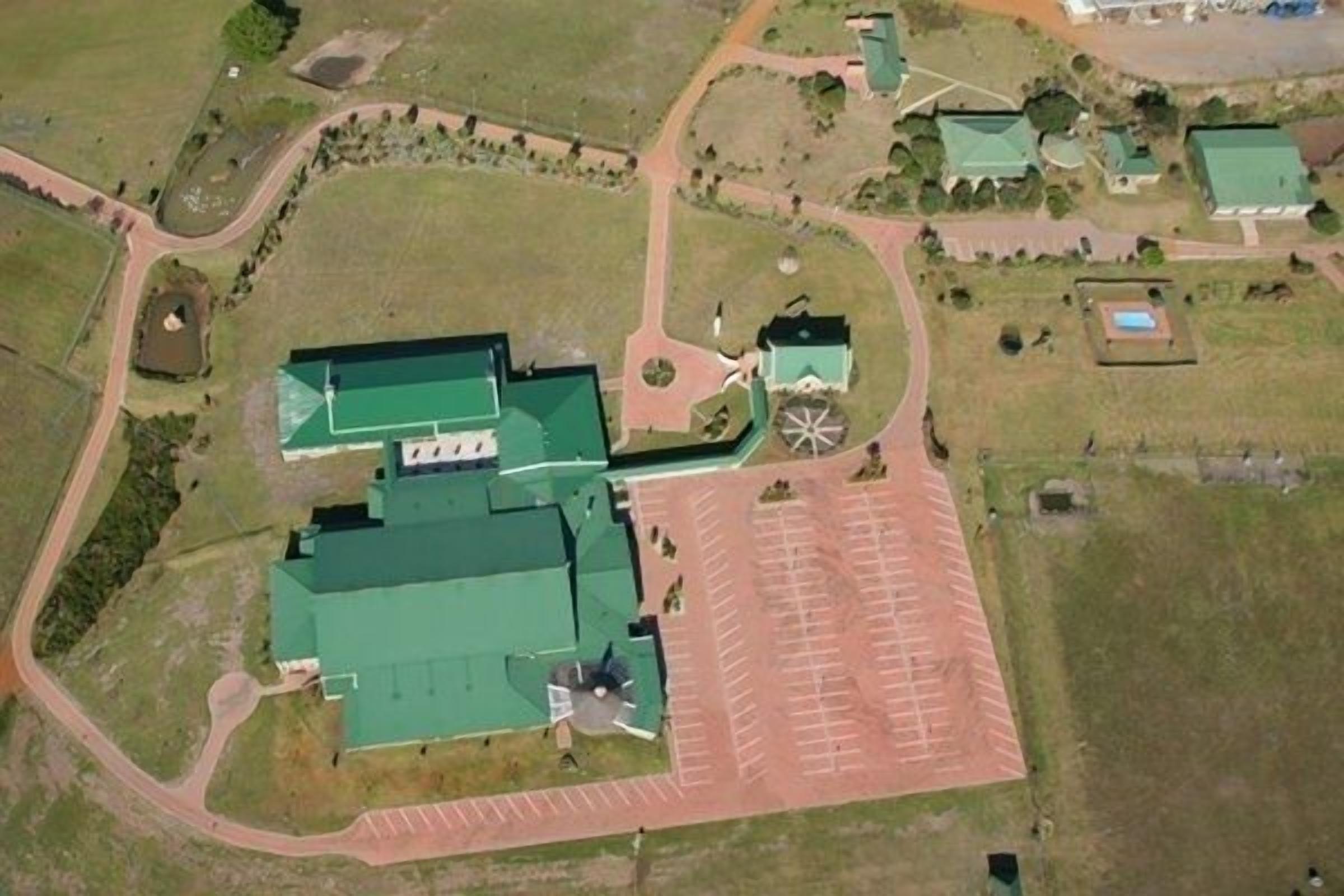 An aerial view of a large building with a green roof, surrounded by parking spaces and a few smaller structures.
